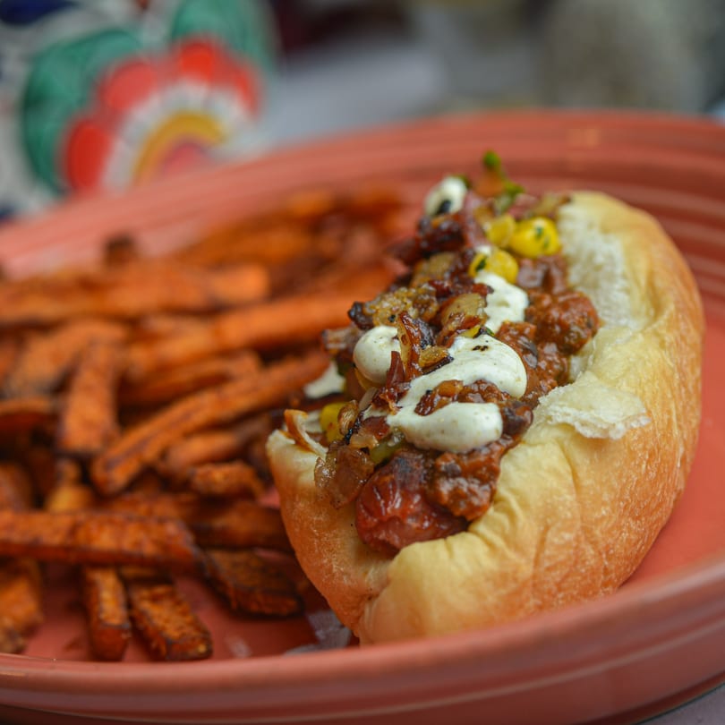 A chili dog with white sauce and caramelized onions, served with sweet potato fries on a red plate.