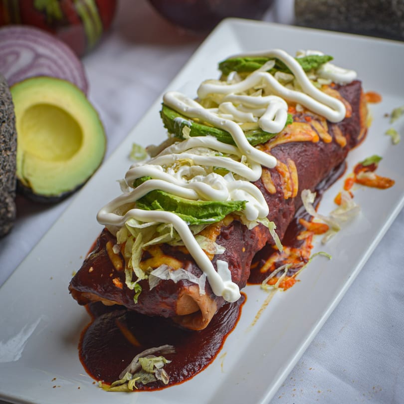 Two enchiladas covered in red sauce, cheese, lettuce, avocado, and sour cream, with avocado in the background.