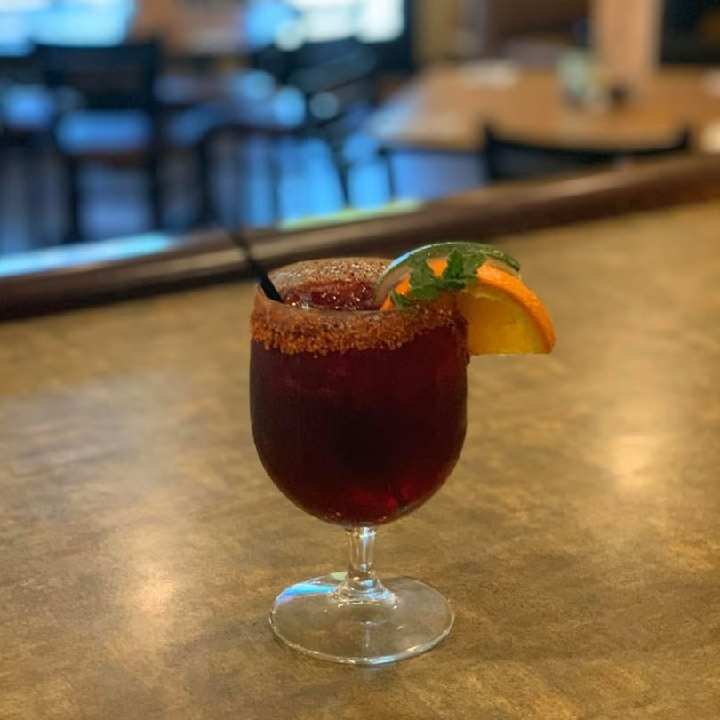 A dark red Mexican drink with a chili-lime rim, orange slice, and mint garnish.