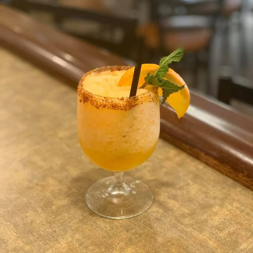 Frozen orange margarita with a chili-lime rim, orange slice, and mint garnish on a bar.