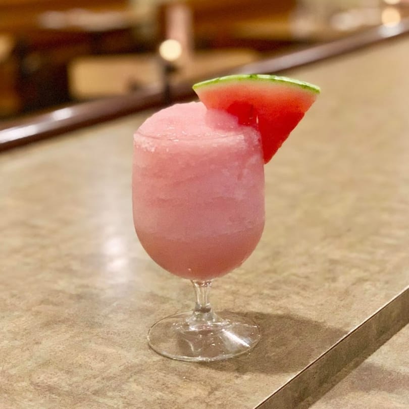 A pink frozen drink, likely a margarita, garnished with a watermelon slice on a bar.
