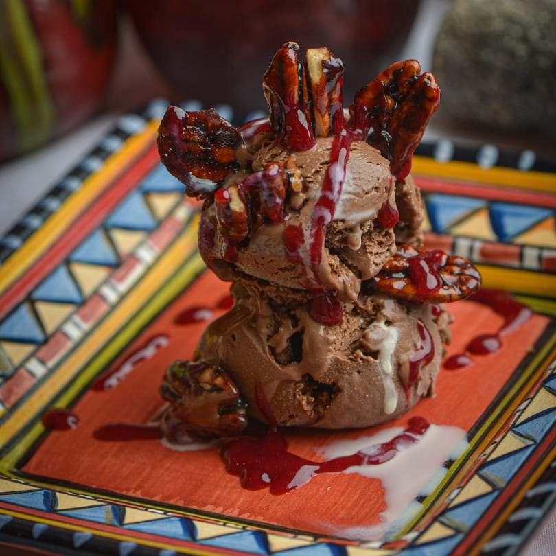 Chocolate ice cream scoops topped with candied pecans and raspberry sauce on a colorful Mexican plate.